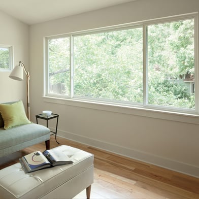 A living room with white walls and wood floors has an expansive view of the tree canopy outside through its three-sash Andersen® 100 Series Gliding Window.